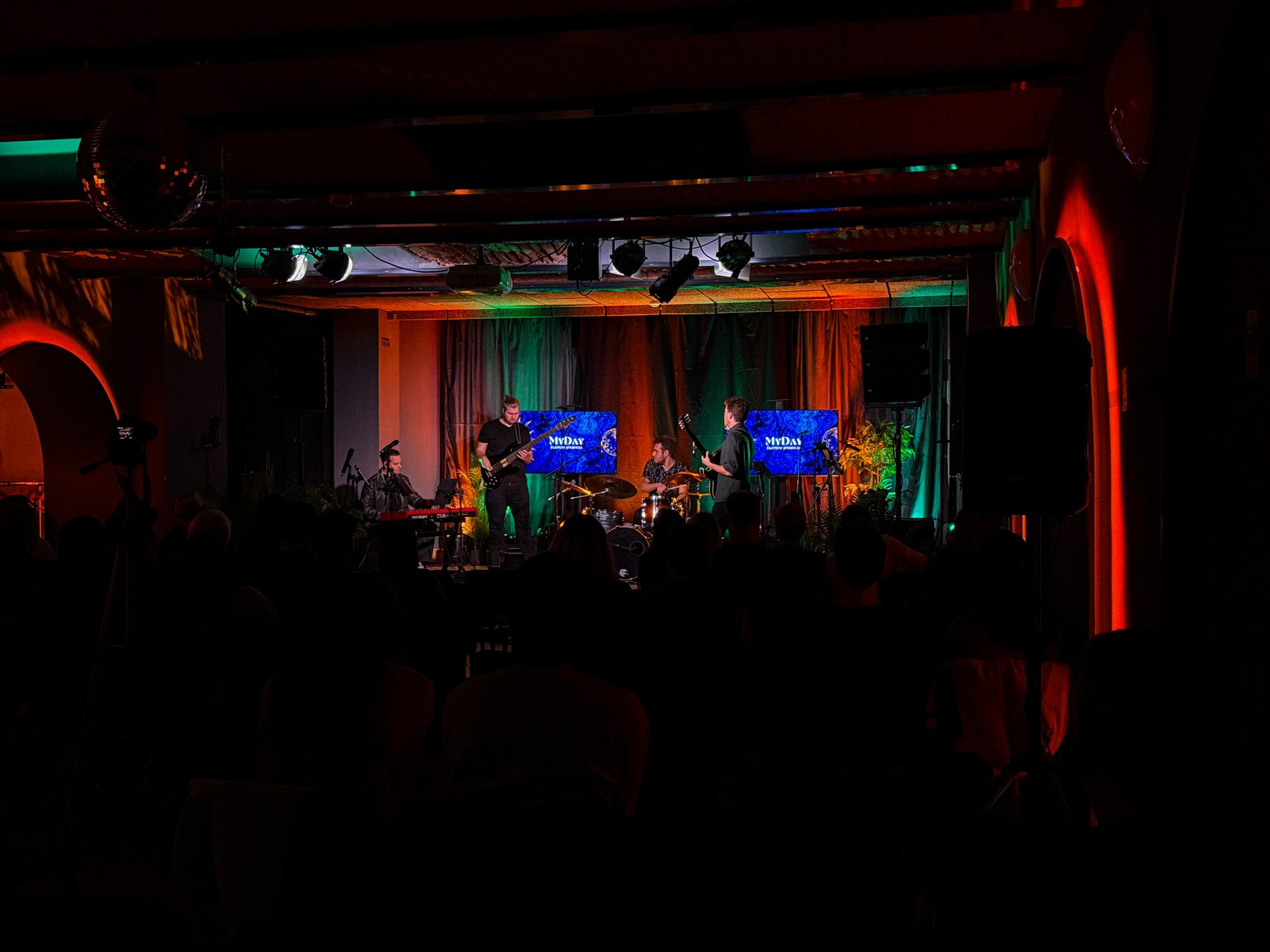 A four-piece band on a stage lit in red and green, audience in silhouette